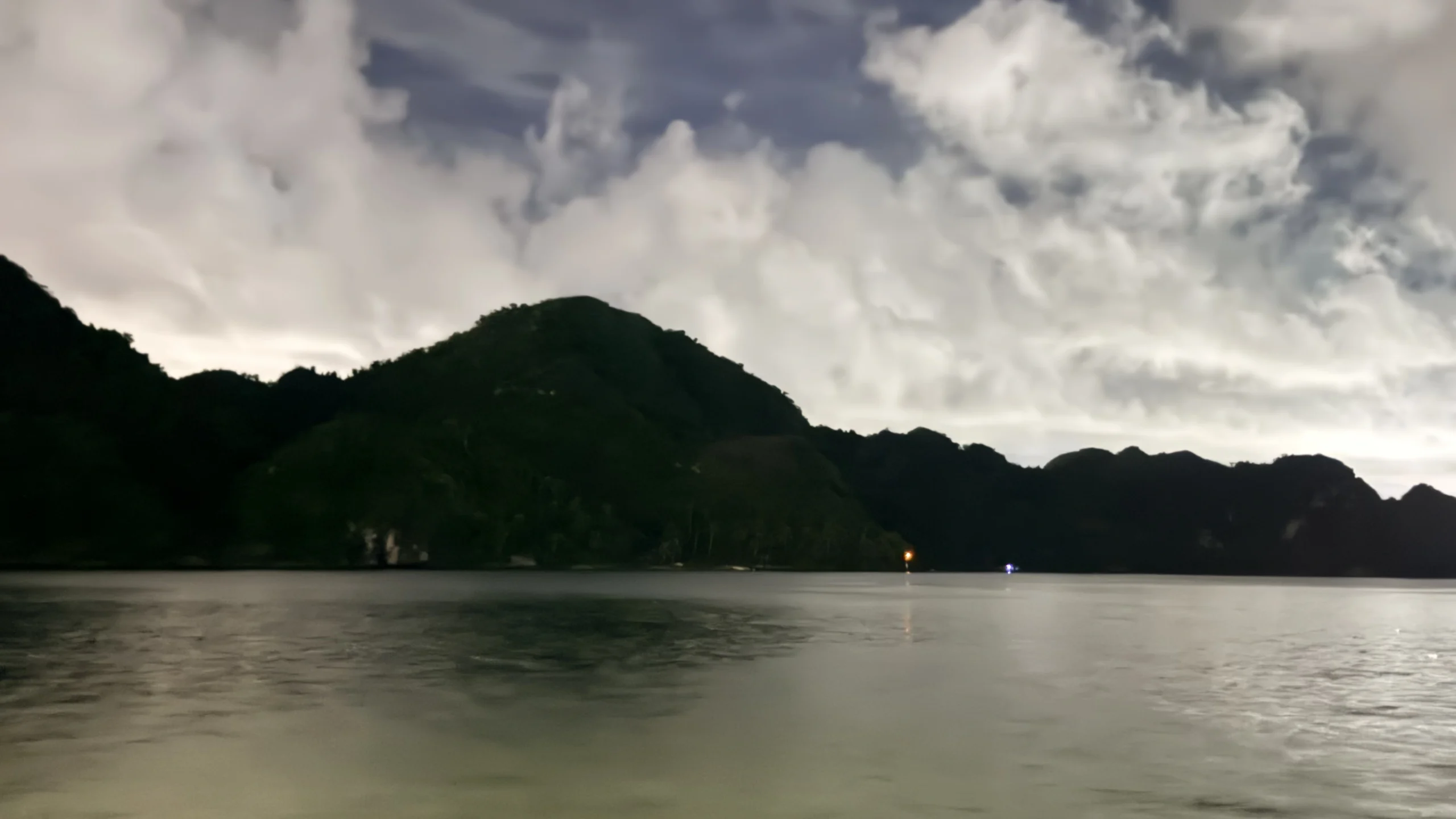 Dark silhouette of limestone mountains across calm bay water under evening clouds in Ha Long Bay
