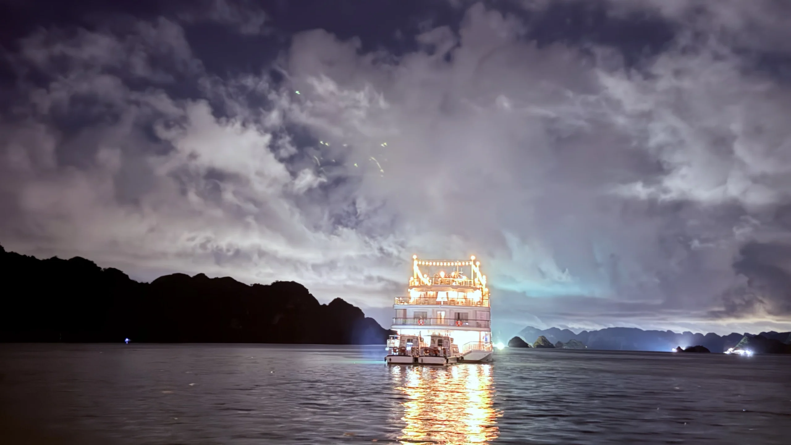 Brightly lit luxury cruise ship anchored near karst mountains at night under a cloudy sky.